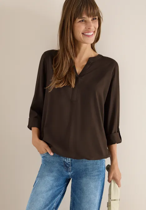 Frau mit brauner Bluse und Jeans lächelt, eine Hand in der Tasche.